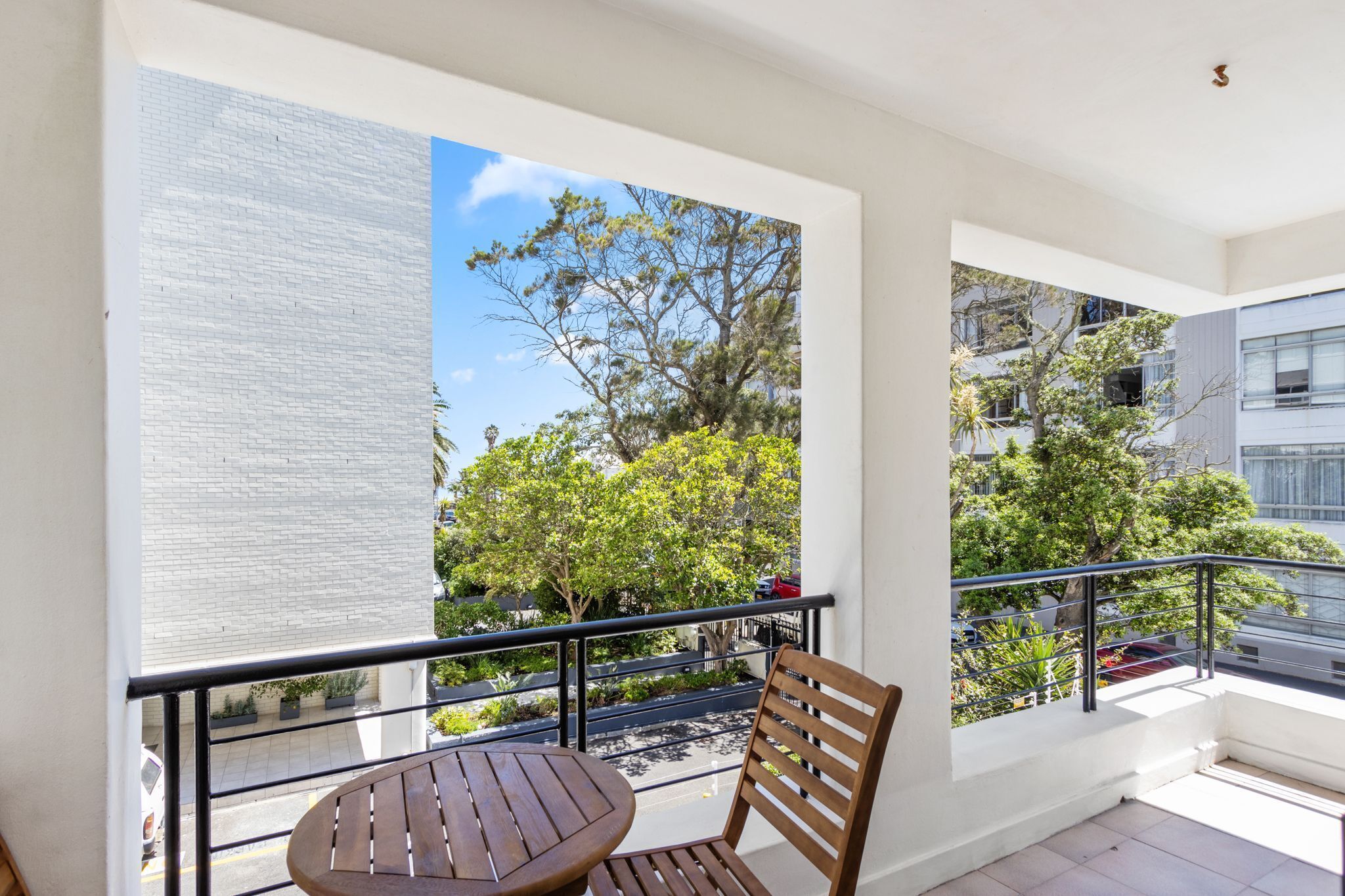 A balcony with a wooden table and chair, a view of trees and buildings, and a clear sky.