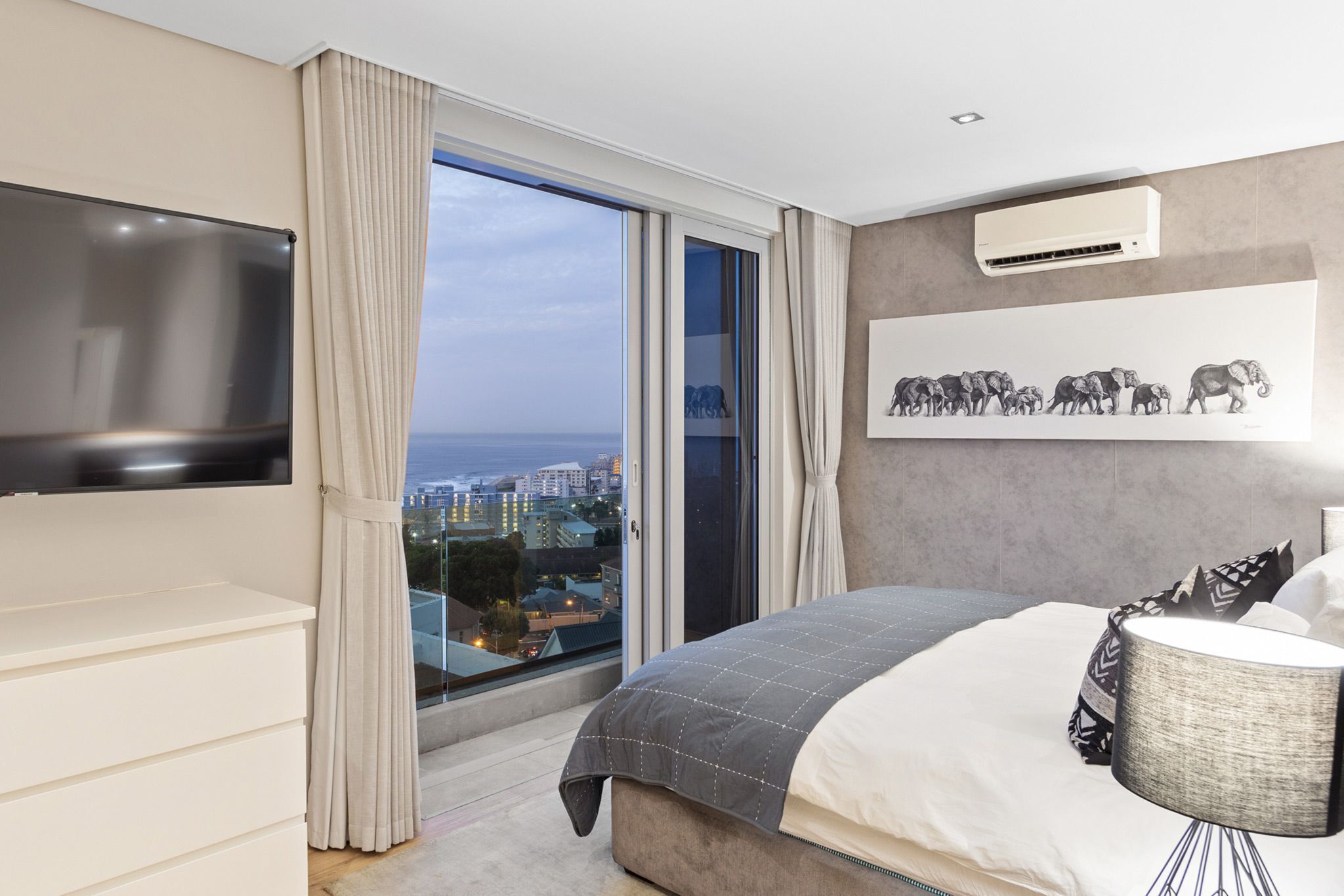 A modern bedroom with a large bed, a flat-screen TV, a city view, and an air conditioning unit.