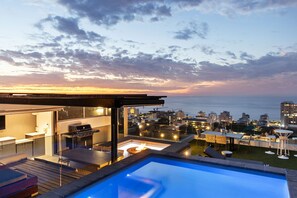 Un espace piscine aménagé sur le toit, avec un barbecue et des sièges, offrant une vue sur le paysage urbain au coucher du soleil.