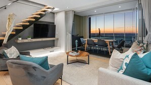 A modern living room with a staircase, a flat-screen TV, and a dining area with a view of the sunset. - Elements on Battery (Cape Town)