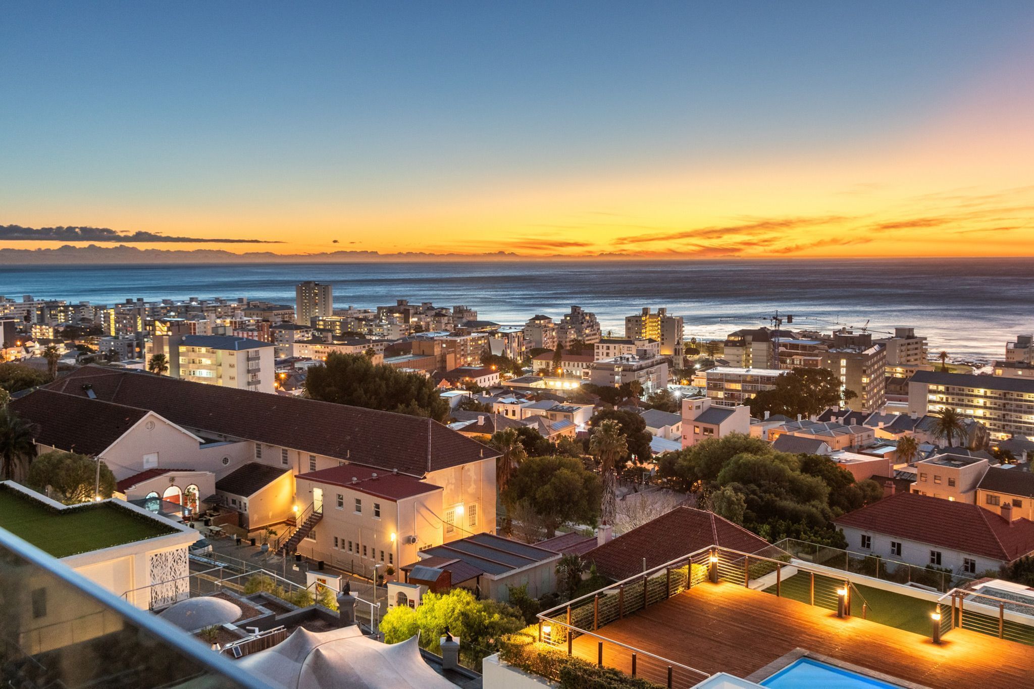 Un paisaje urbano costero al atardecer con edificios, una piscina y vistas al océano.