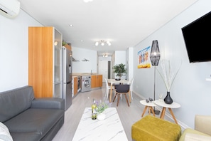 Un salon moderne comprenant un canapé, une table à manger avec des chaises, un téléviseur fixé au mur et un coin cuisine équipé d’appareils électroménagers.