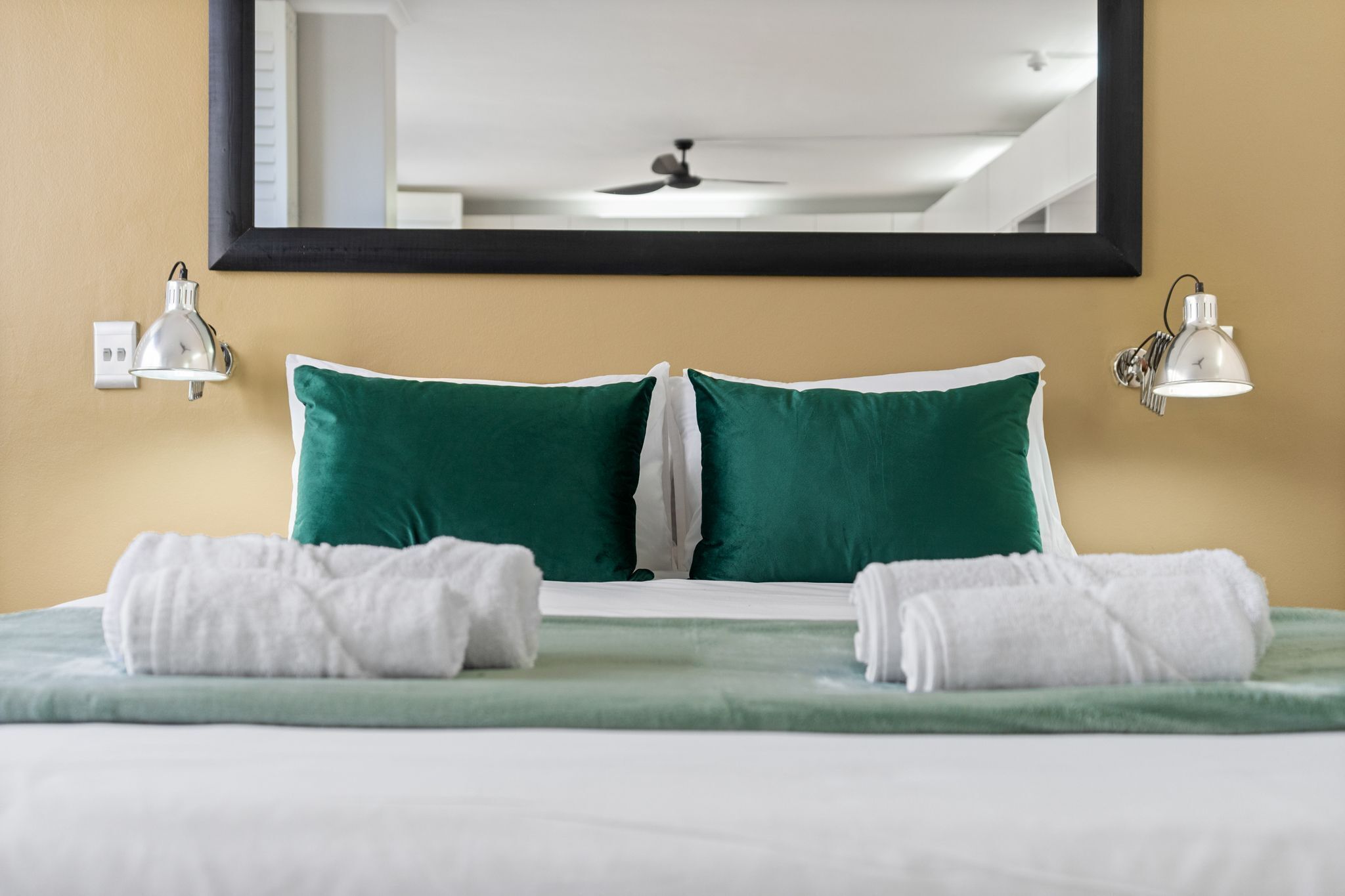 Habitación de hotel con cama, dos almohadas verdes, dos toallas blancas, ventilador de techo y dos lámparas de pared.