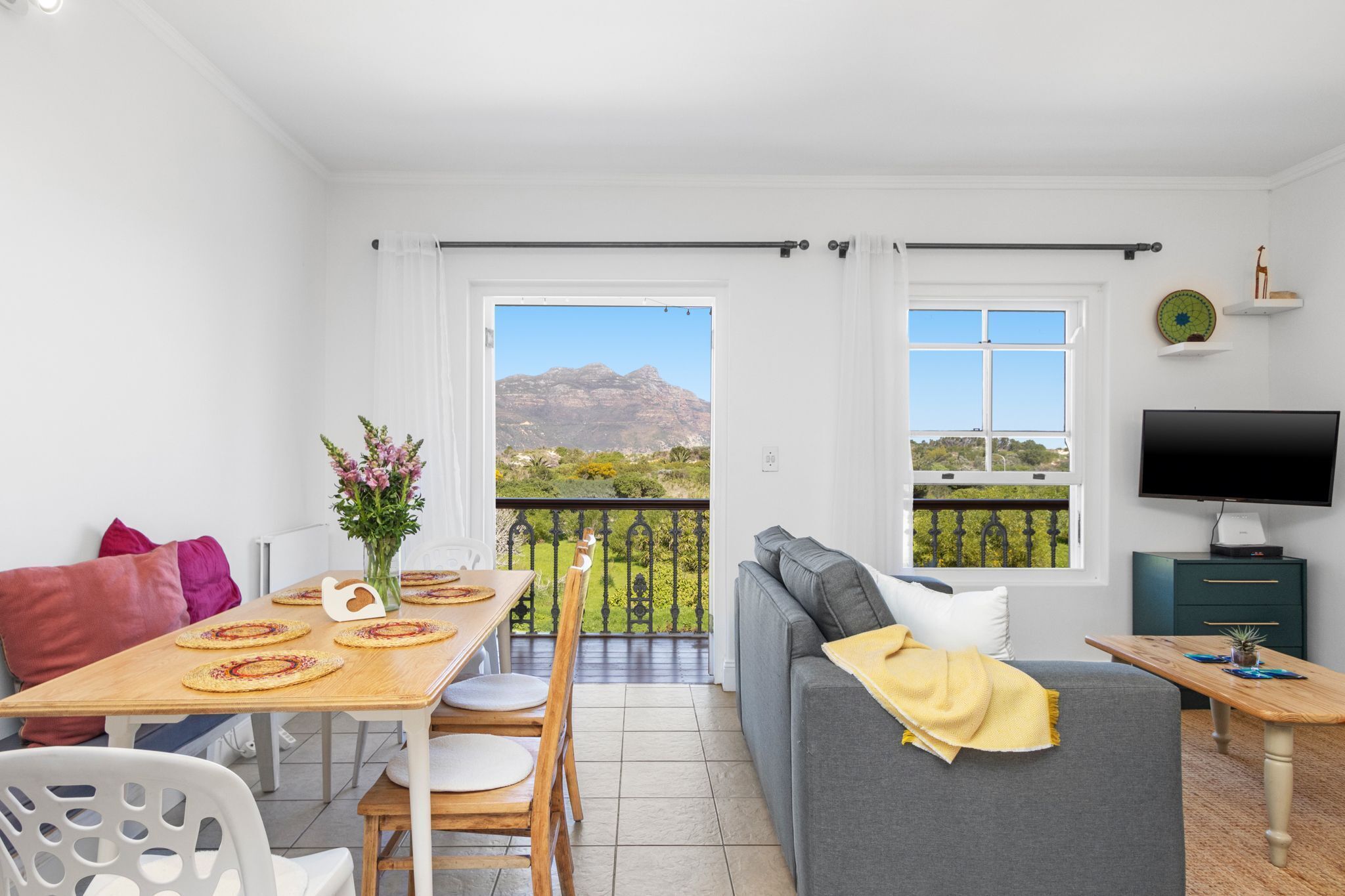 En moderne stue med balkon, spiseplads med et bord dækket til to personer, og et fjernsyn på et stativ.