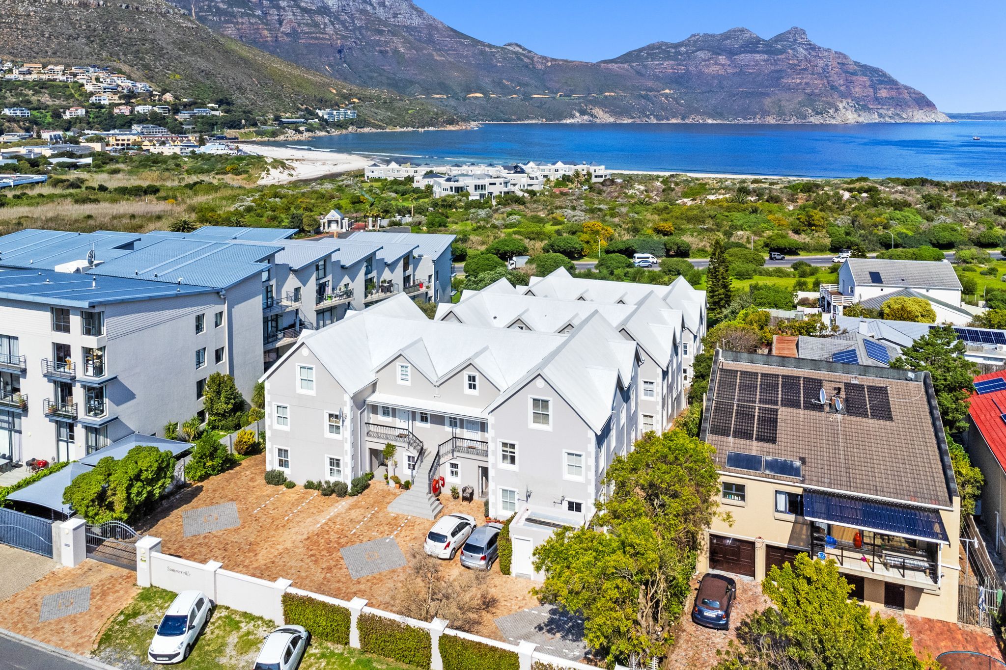 A residential area with modern houses, a large building with a staircase, and a beach in the background.