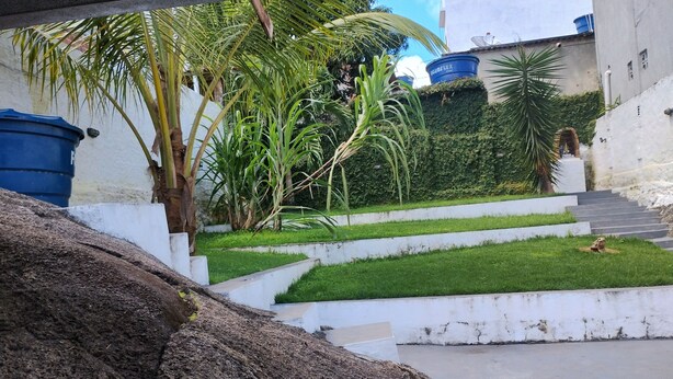 Un área ajardinada con un sendero de concreto, palmeras y un depósito azul con la etiqueta 'KUNDFLEX'.