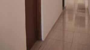 Un pasillo con una puerta de madera a la izquierda, una pared blanca y un suelo de baldosas.