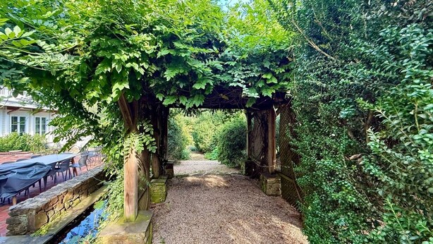 Un sendero cubierto con superficie de grava, rodeado de exuberante follaje y una estructura de pérgola de madera.