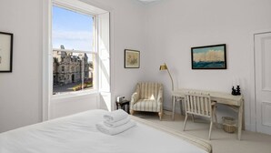 A bedroom with a large window, a bed, a desk, a chair, and two framed pictures.