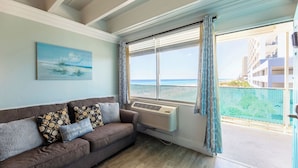 A room with a large window overlooking a beach, a sofa with patterned cushions, and a painting of a beach scene. - 308 Beachfront Condo with Pool Sunset Views (panama city beach)