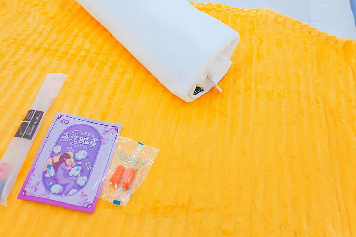 A bed with white linens and a peach pillow, a rolled white blanket, and a yellow blanket on a textured orange surface.