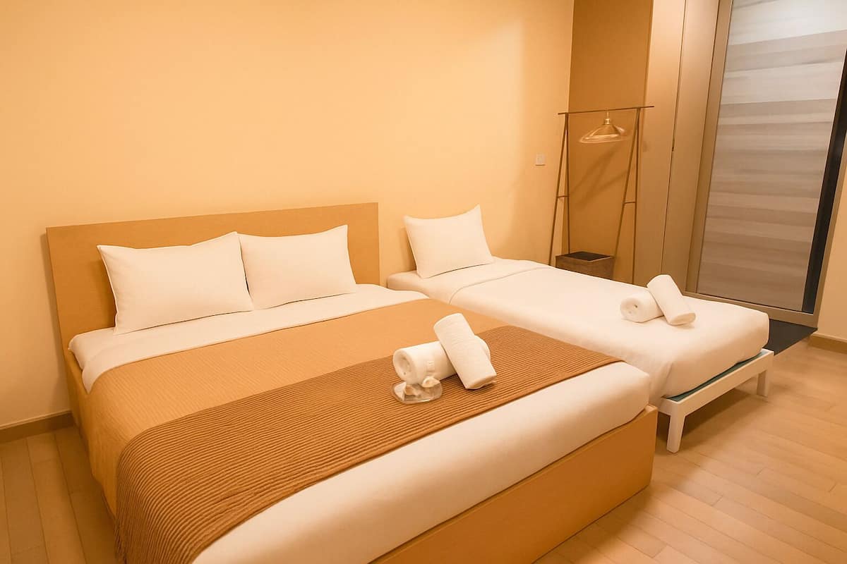A double room with two beds, a wooden headboard, a large window with blinds, and a bedside table with a lamp.