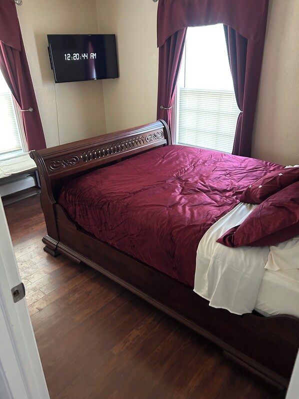 Una cama bien hecha con cabecera de madera, un televisor montado en la pared y una ventana con cortinas.