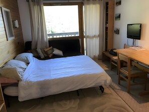 Een slaapkamer met een bed, een bureau, een stoel, een televisie en een raam met gordijnen.
