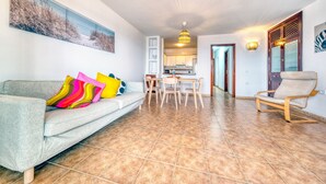 A living room with a sofa, colorful pillows, a dining table with chairs, a wooden chair, and a kitchen in the background. - El Charcón A • 10 meters to the beach • LVH (Arrieta)