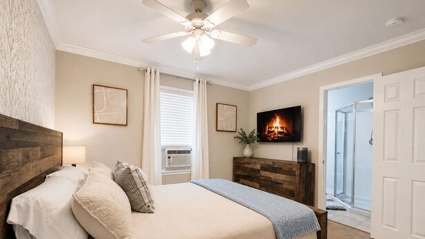 Un dormitorio con cabecera de madera, cama con ropa de cama blanca, ventilador de techo, chimenea y televisor.