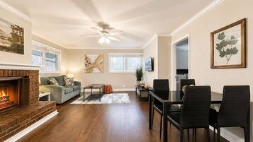 A living room with a fireplace, a ceiling fan, a dining area with chairs, and artwork on the walls.