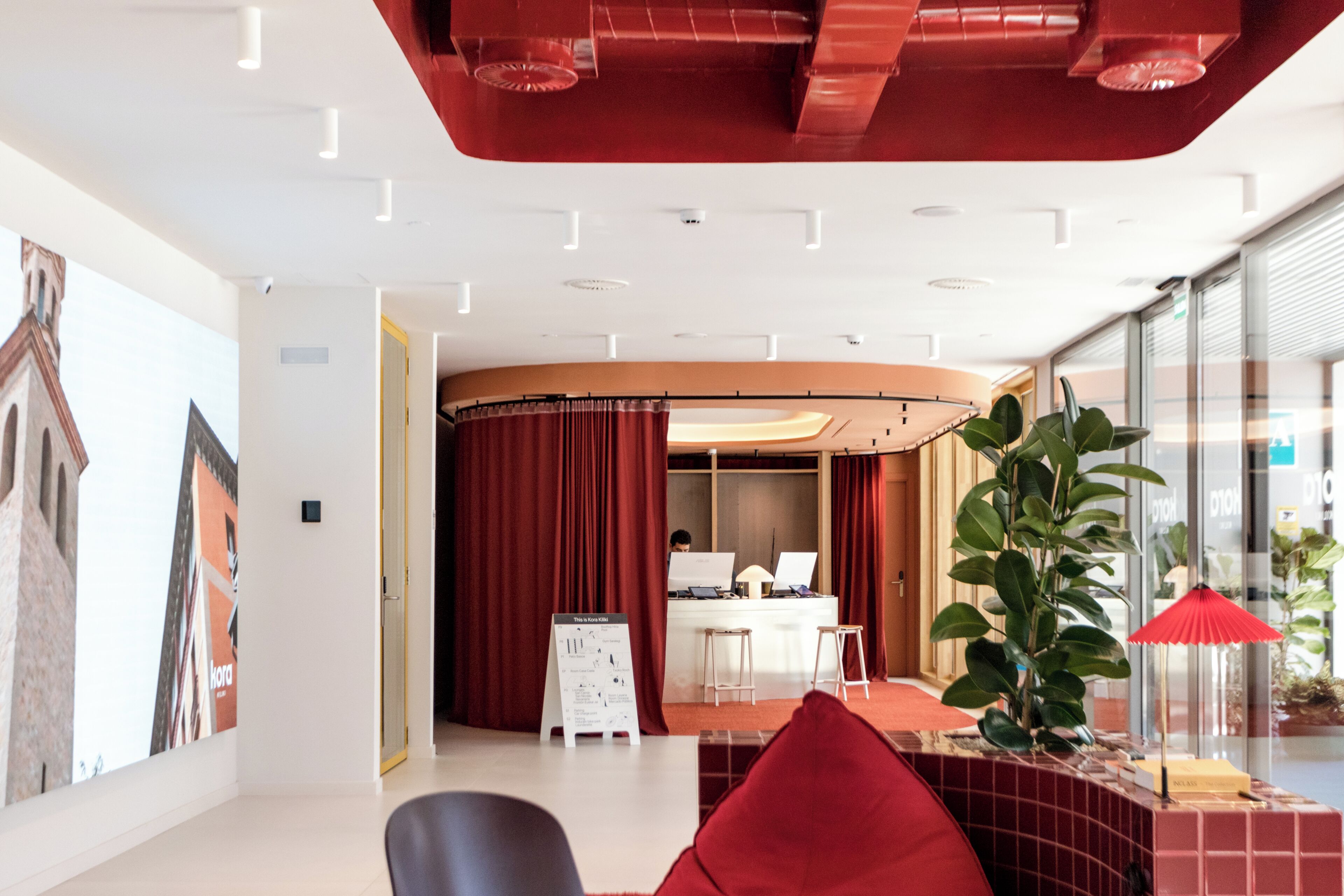Un moderno vestíbulo de hotel con una recepción, un área de descanso con cojines rojos y una planta grande.