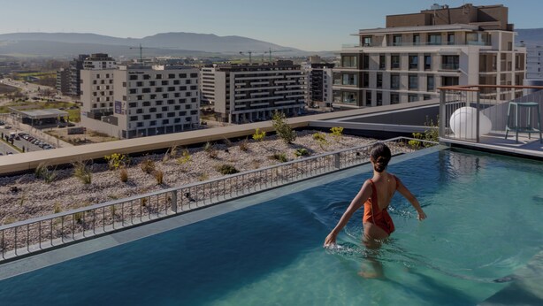 A swimming pool with a person in an orange swimsuit, surrounded by modern buildings and a clear sky. - KORA KILIKI (Pamplona)
