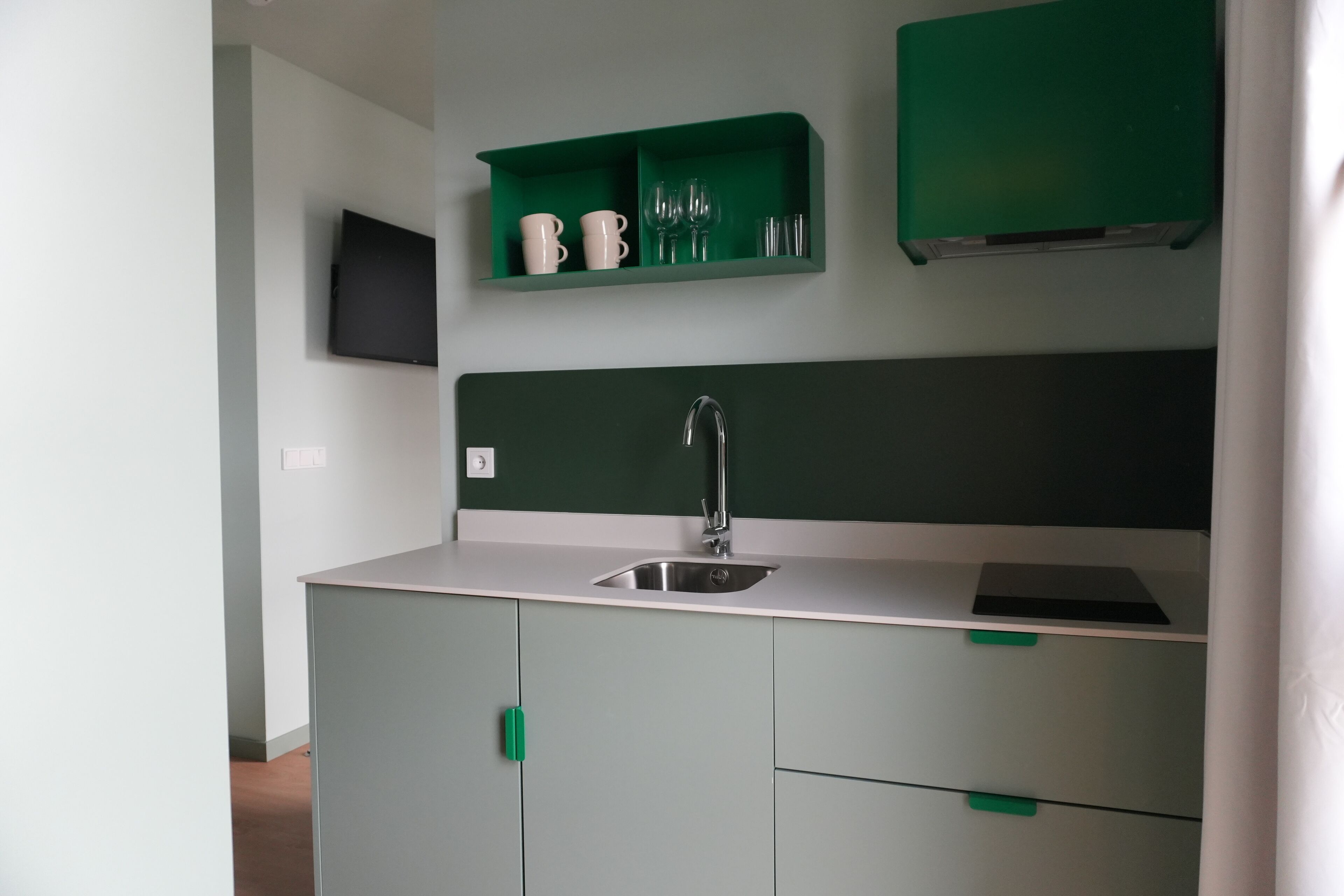 A kitchen with a sink, green cabinets, and a green shelf with cups and glasses.