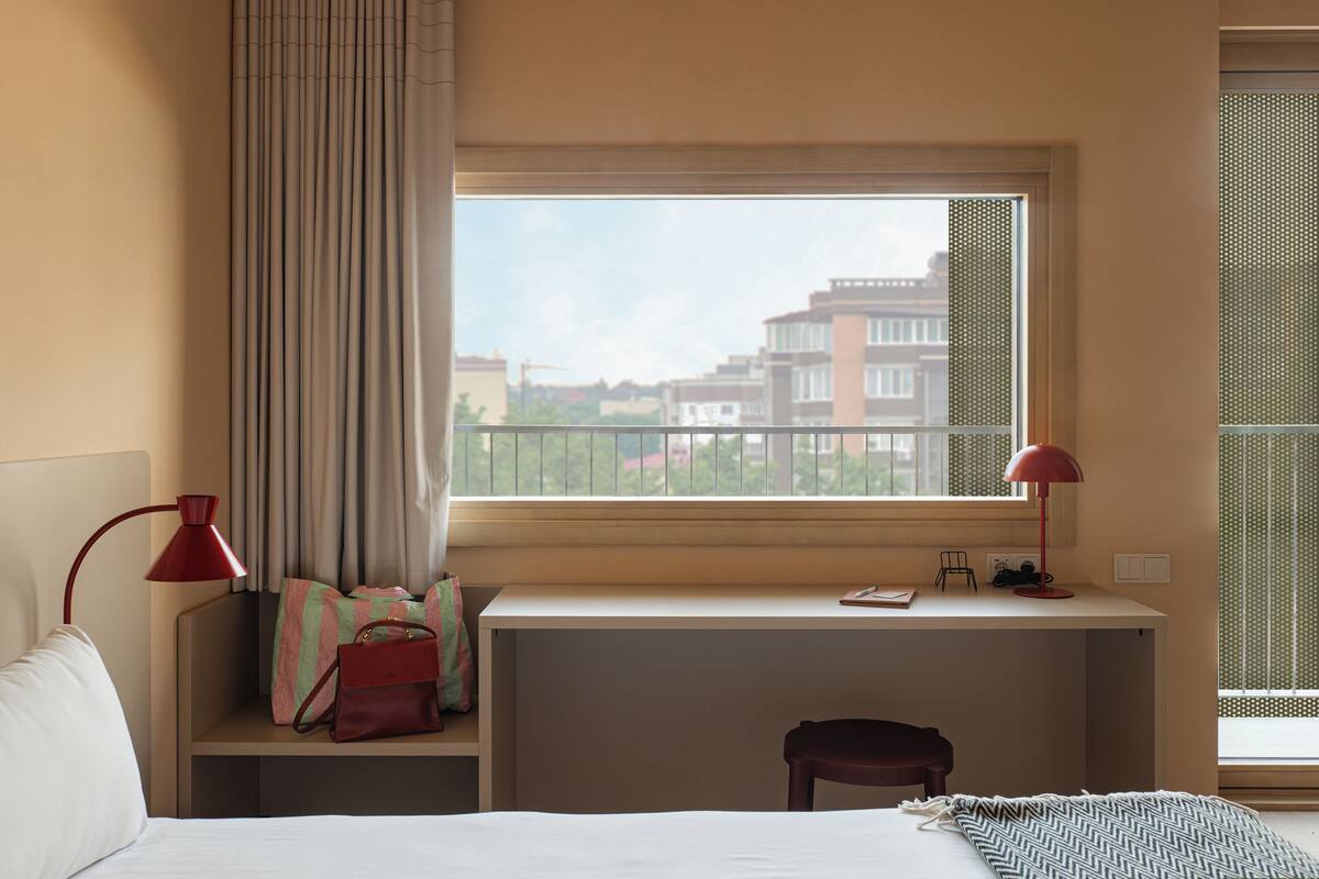 A hotel room with a bed, a desk with a chair, a window with a view of buildings, and a red lamp.