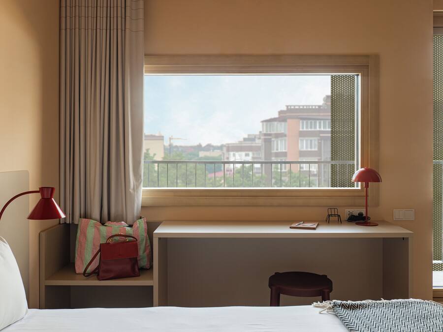 Habitación de hotel con cama, escritorio y silla, ventana con vista a edificios y una lámpara roja.
