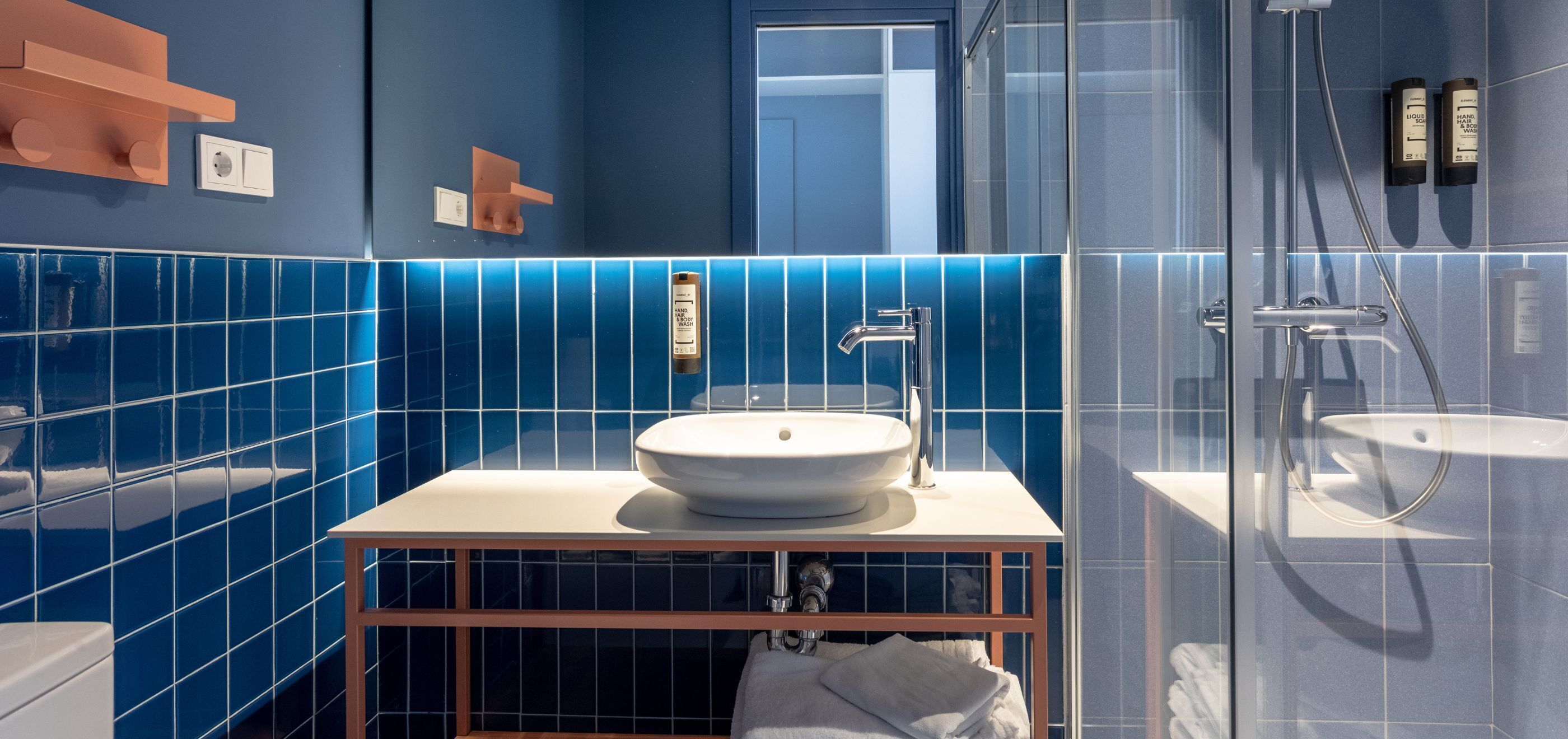 Une salle de bain moderne avec un lavabo blanc, des murs recouverts de carreaux bleus, une douche en verre et un meuble-lavabo en bois.