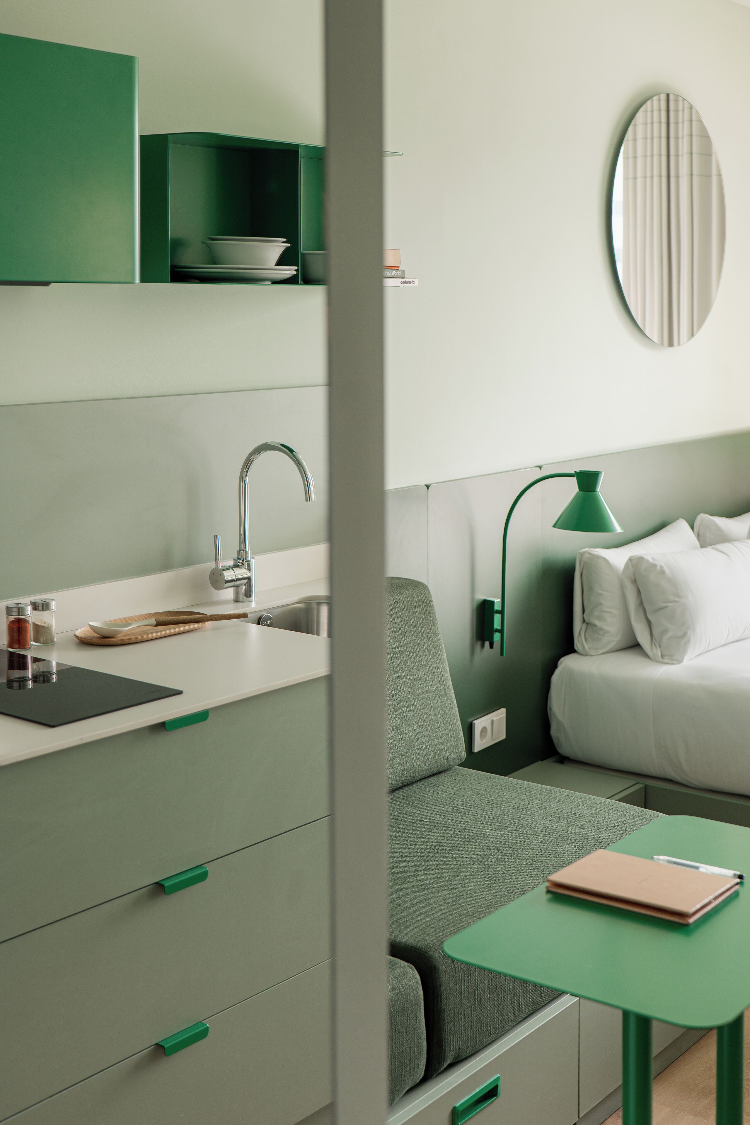 Una habitación de hotel moderna con kitchenette, un sofá verde, una mesita y una cama con ropa de cama blanca.
