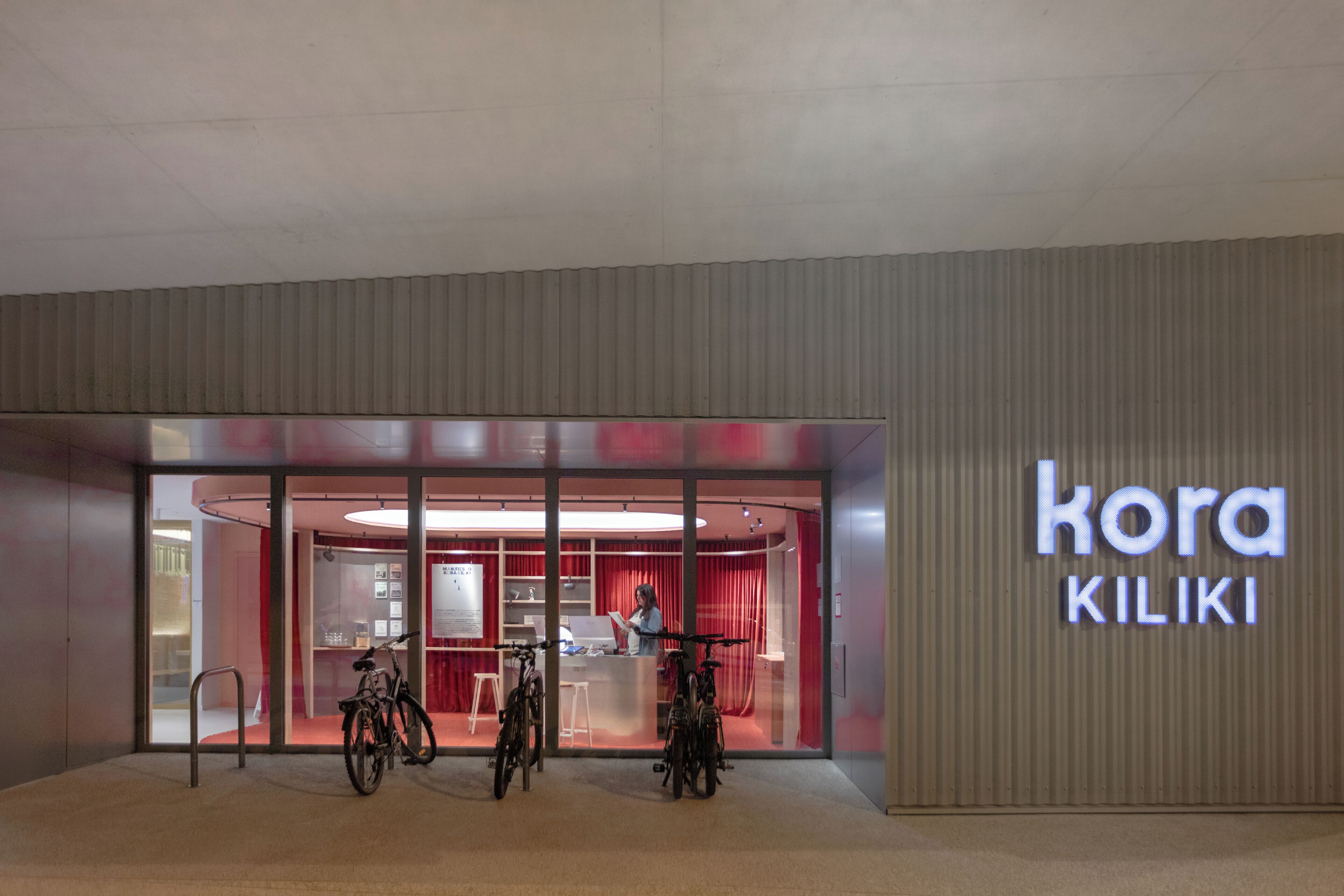 Une entrée moderne, avec une façade en verre, des vélos garés à l’extérieur et une enseigne indiquant « kora KILIKI ».
