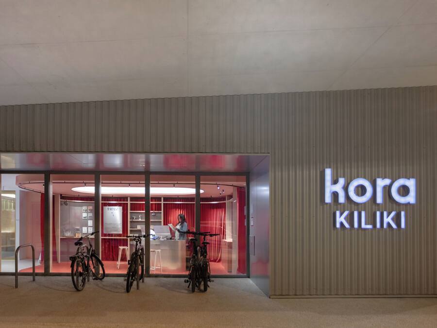 Une entrée moderne, avec une façade en verre, des vélos garés à l’extérieur et une enseigne indiquant « kora KILIKI ».