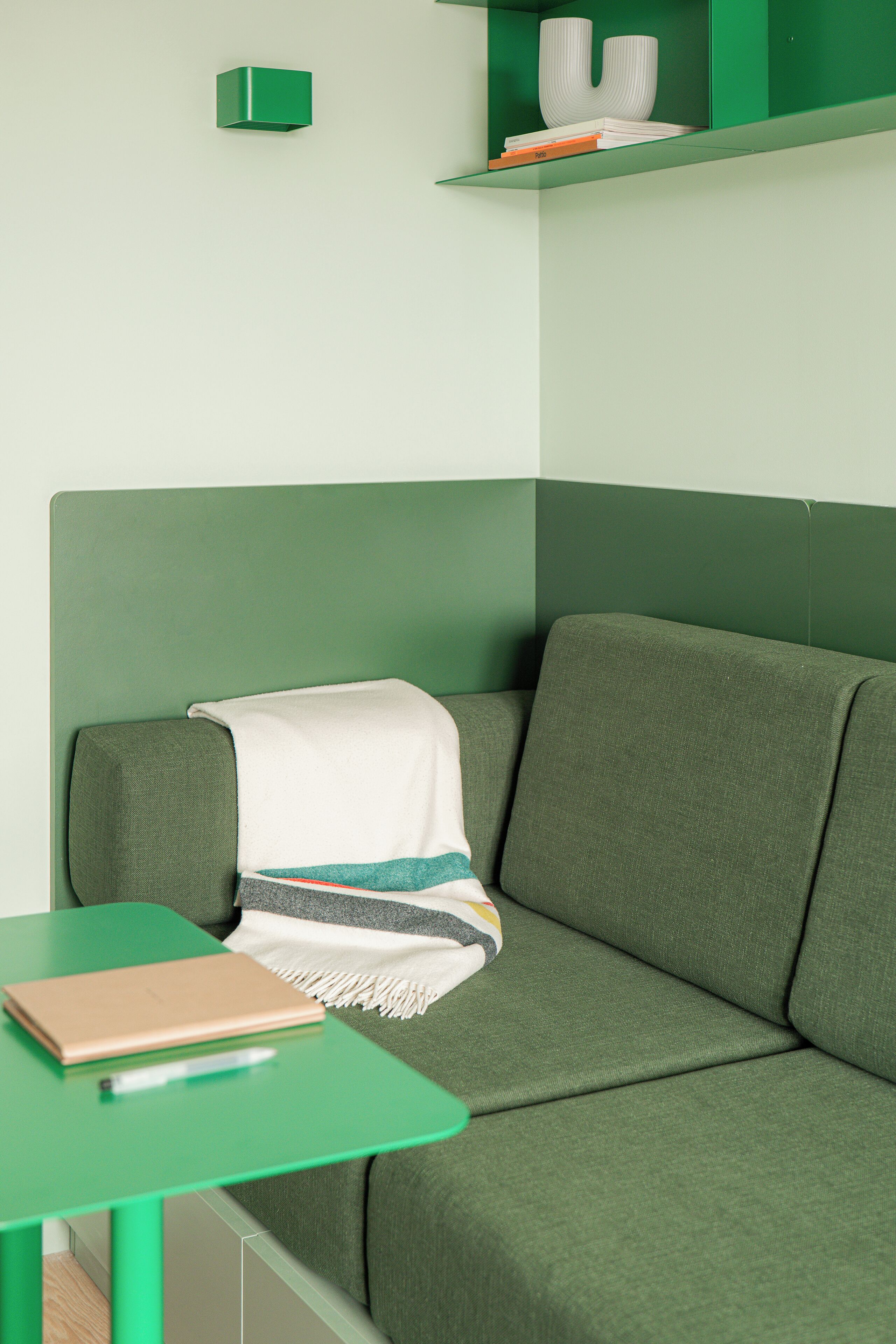 Un canapé vert avec un plaid blanc, une table verte sur laquelle se trouve un carnet, et une étagère verte ornée d’un vase blanc et de livres.