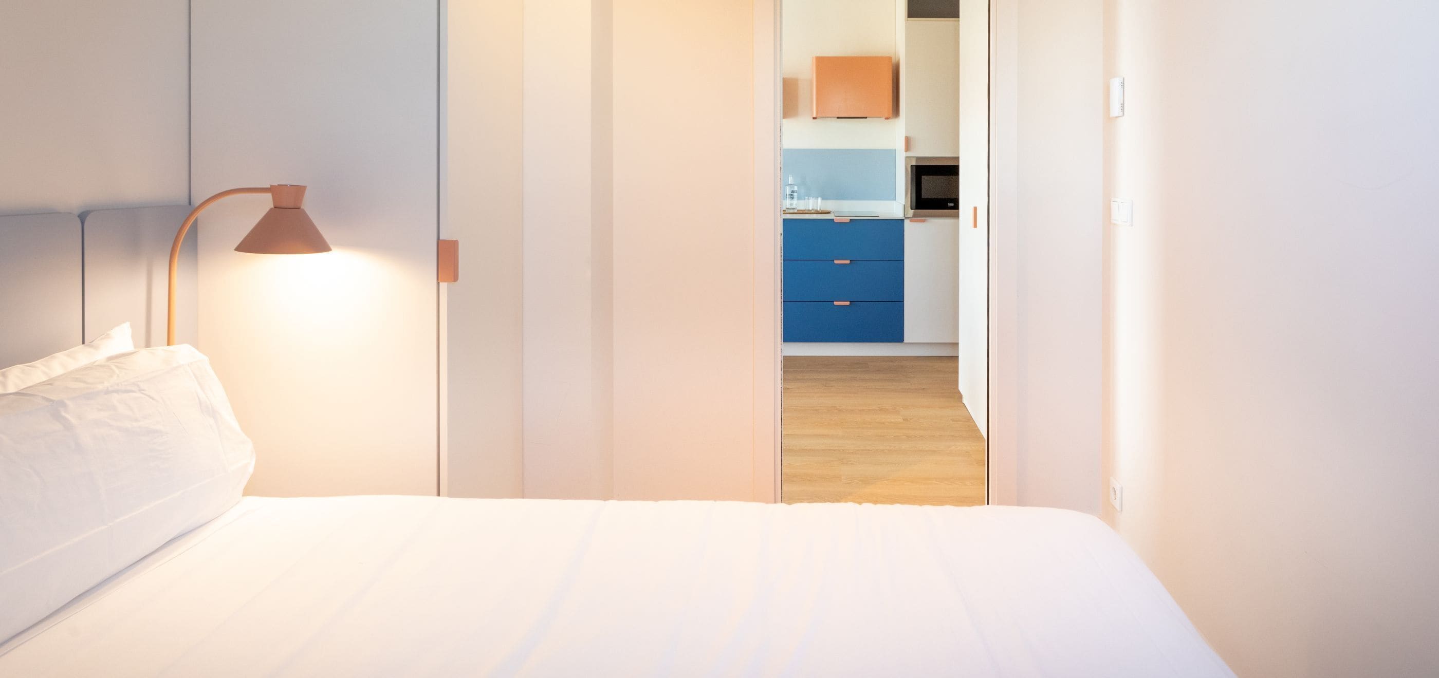 A bedroom with a bed, a bedside lamp, and a view into a kitchen area with cabinets and a microwave.