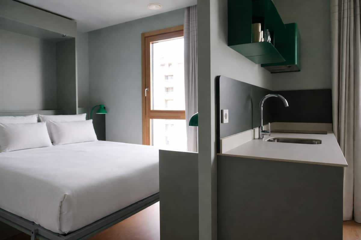 A hotel room with a bed, a small kitchenette, a window with curtains, and a view of a building outside.