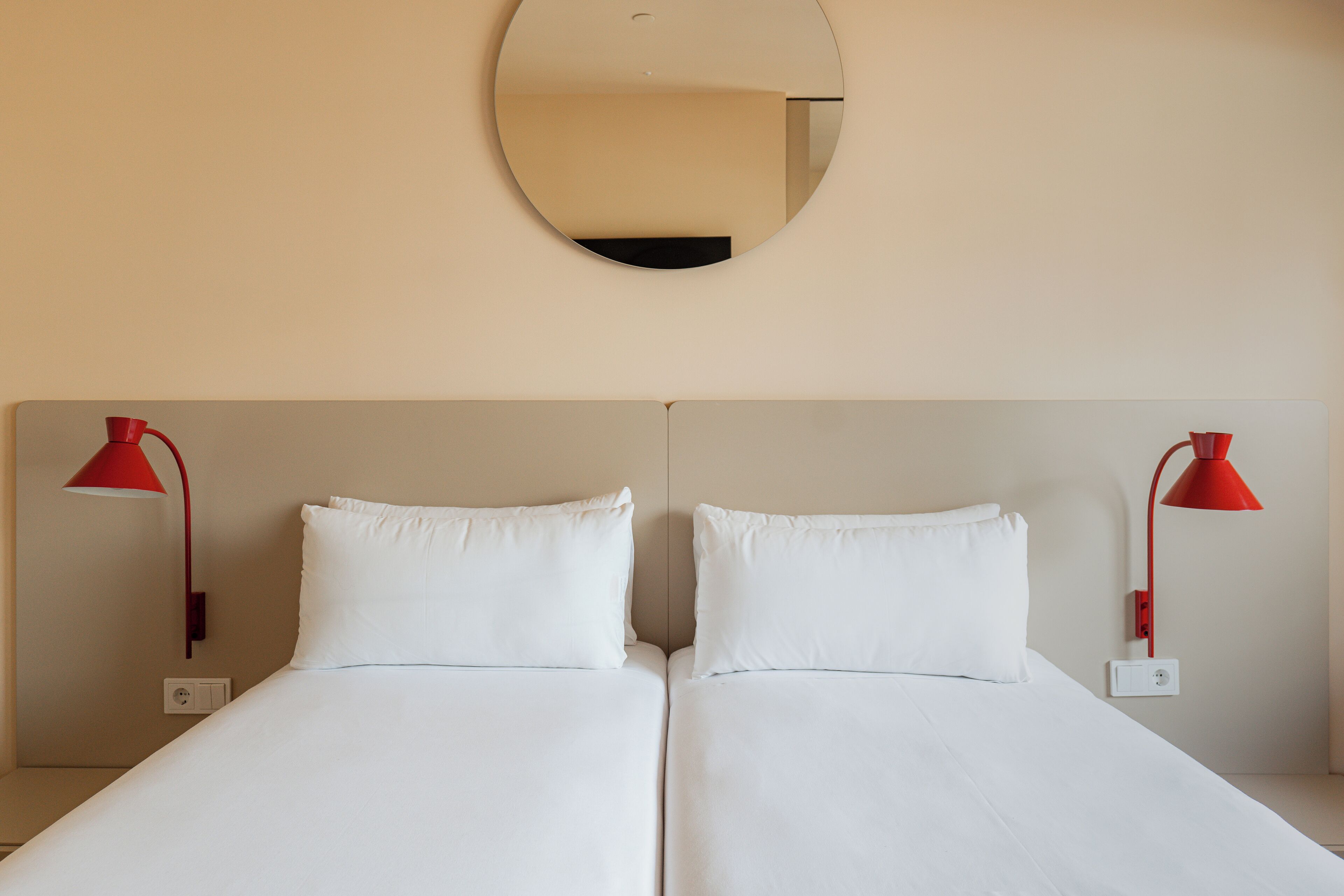 A double bed with white bedding, two red bedside lamps, and a round mirror on the wall.