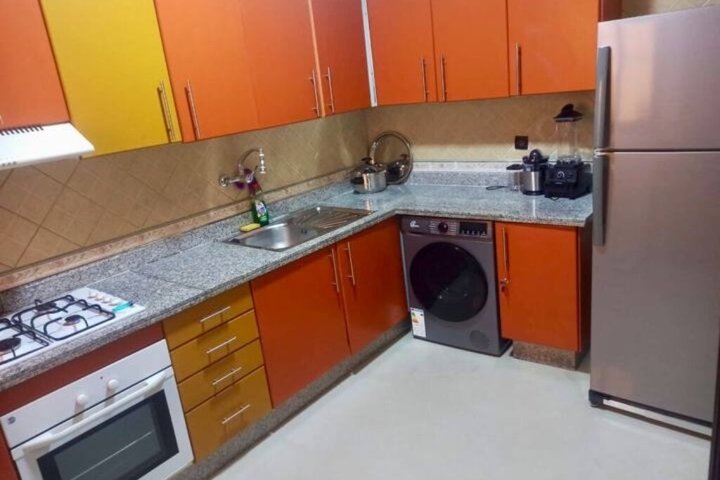 Une cuisine avec des meubles de couleur orange, une cuisinière à gaz, un lave-linge et un réfrigérateur.