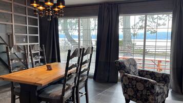 Een eetgedeelte met een houten tafel en stoelen, een kroonluchter en uitzicht op een steiger en bomen door grote ramen.