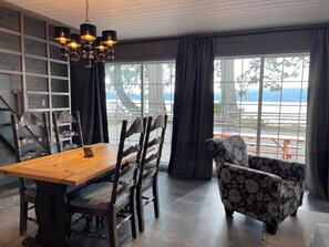 Un espace repas avec une table et des chaises en bois, un lustre et une vue sur un quai et des arbres, visible à travers de grandes fenêtres.