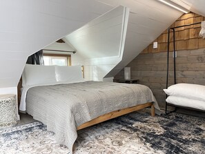 Une chambre avec un plafond en pente, un lit recouvert d’une couette grise, une tête de lit en bois, une table de chevet, une lampe et une fenêtre avec des rideaux.