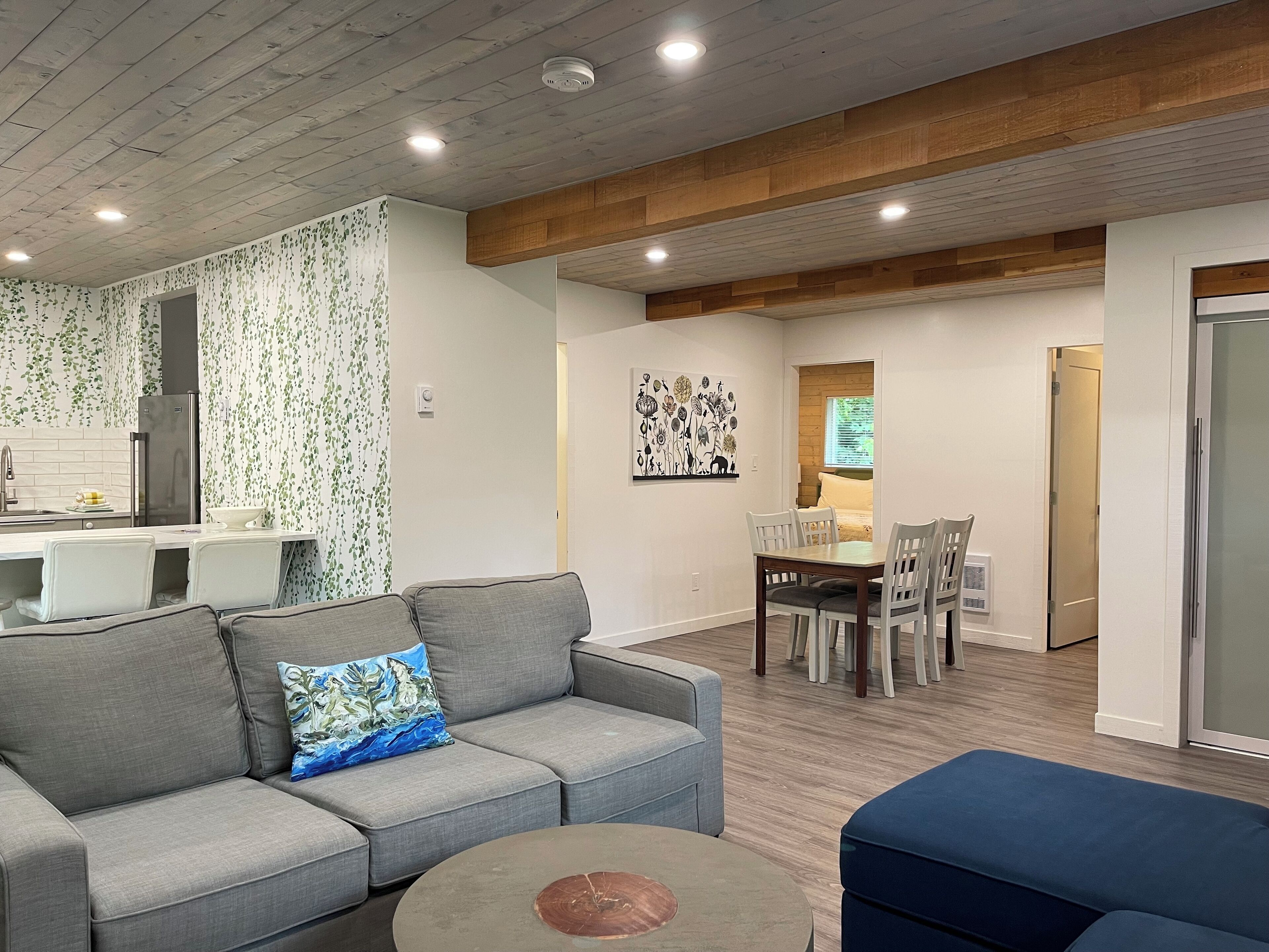 A modern living room with a grey sofa, a blue ottoman, and a dining area with a wooden table and chairs.
