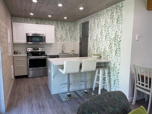A compact kitchen with white cabinets, a microwave, an oven, and a sink. There is a small bar area with stools and a patterned cushion.