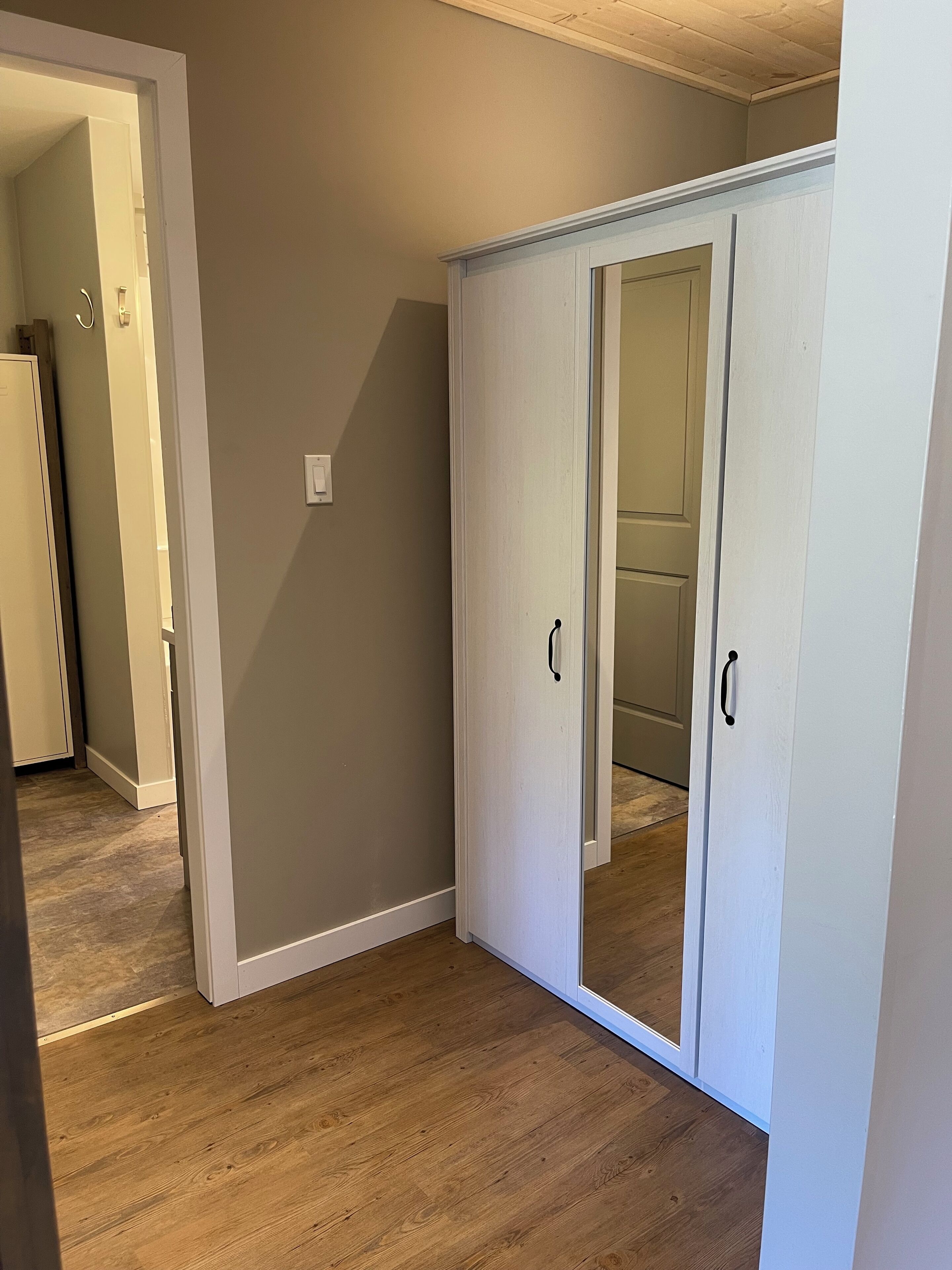 A room with a wooden floor, a white wardrobe with two doors, and a light switch on the wall.
