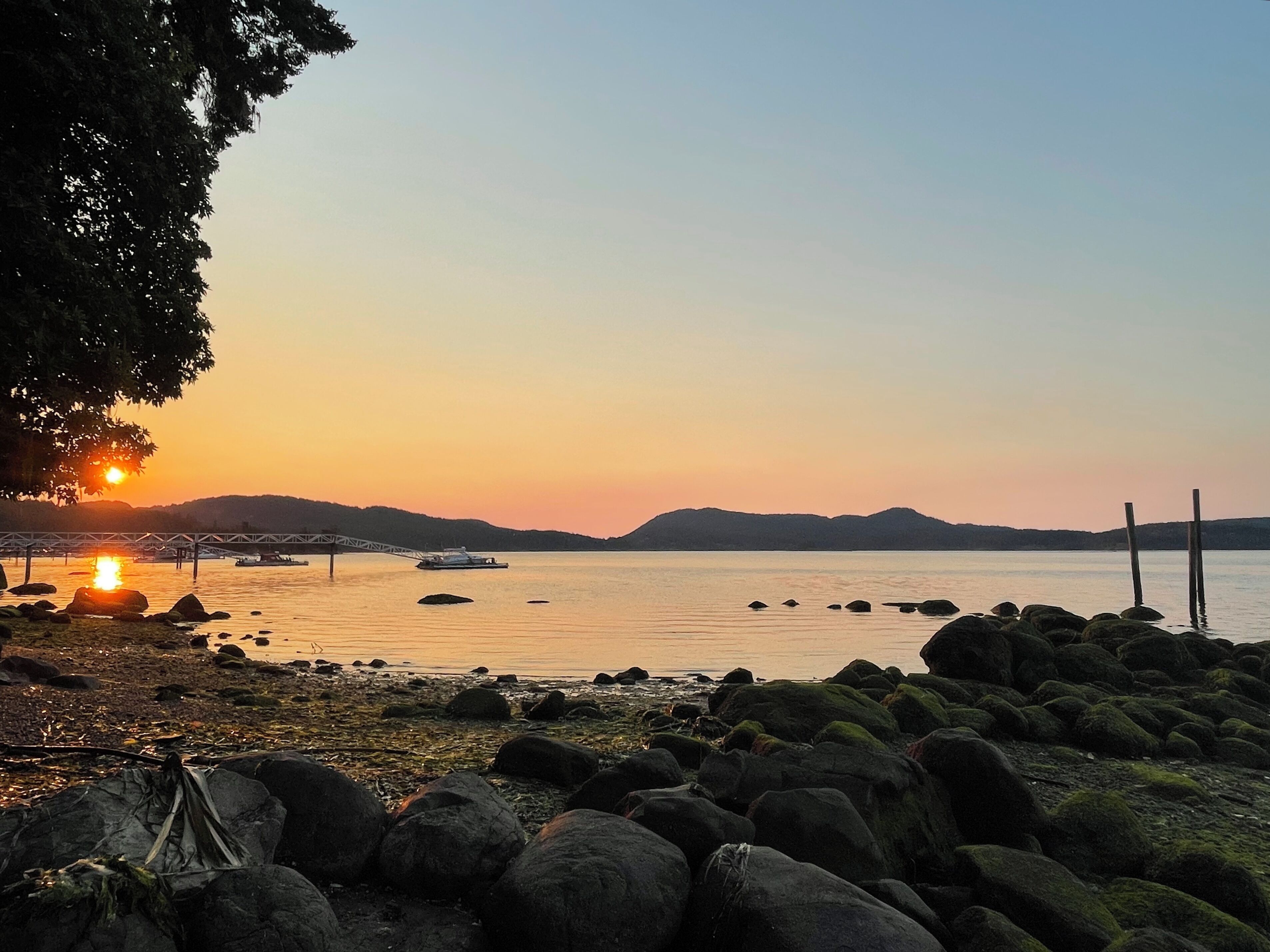 A rocky shoreline with a sunset over a calm body of water, a bridge, and a boat.