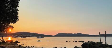 A rocky shoreline with a sunset over a calm body of water, a bridge, and a boat.
