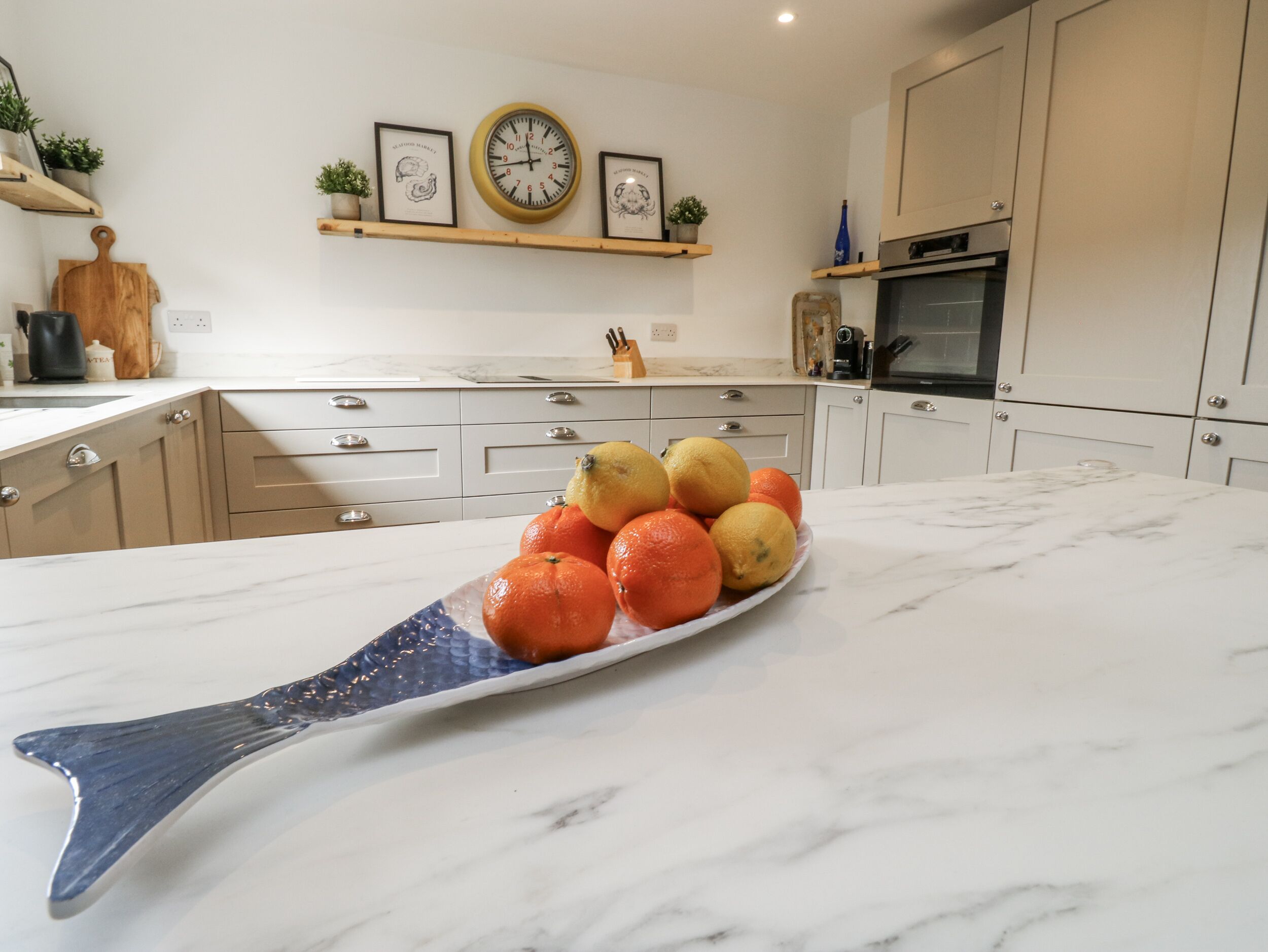 A kitchen with a marble countertop, a clock, and a bowl of oranges.