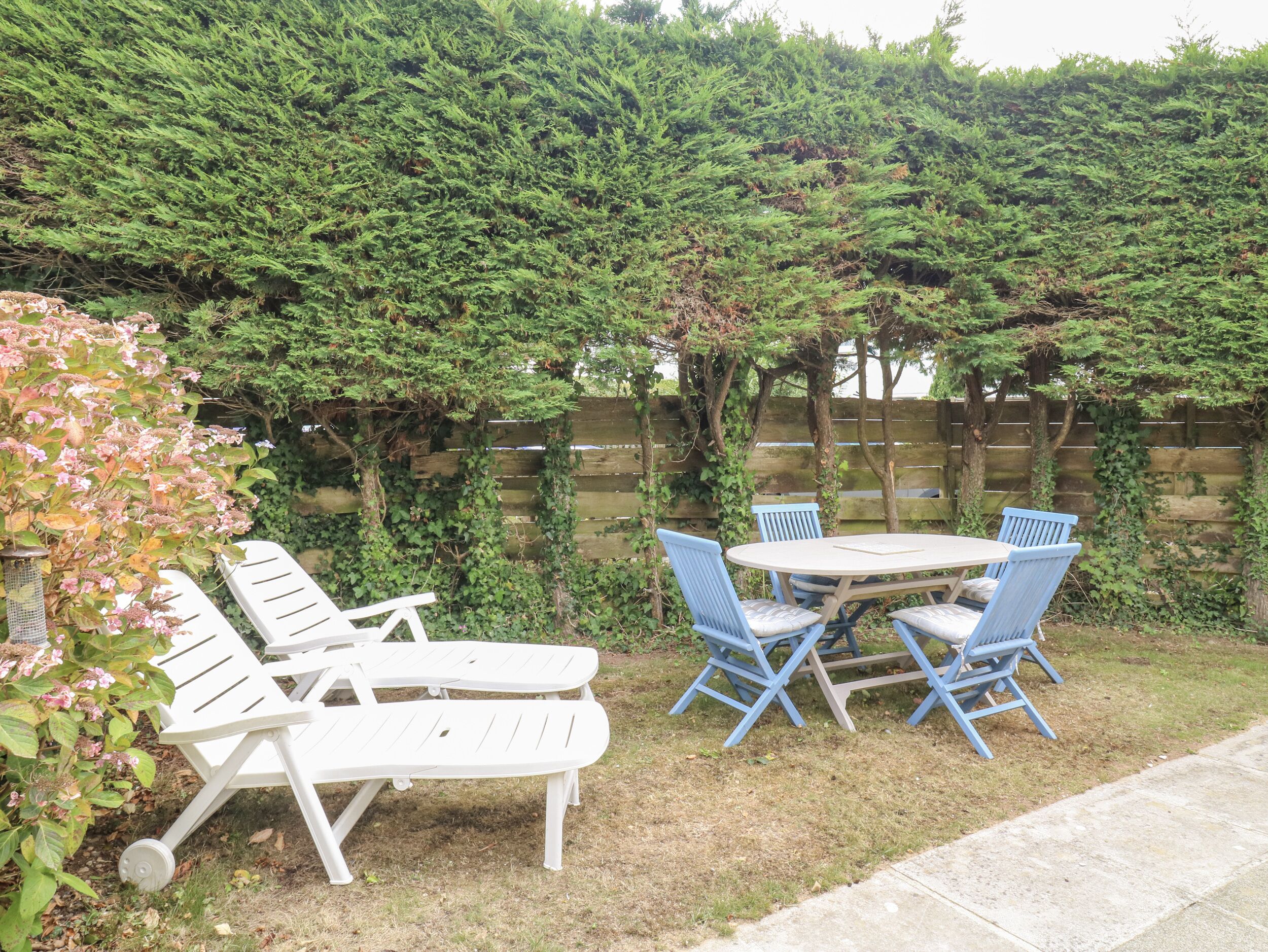 Une terrasse avec une table ronde et quatre chaises bleues, entourée d’une clôture en bois et de verdure.