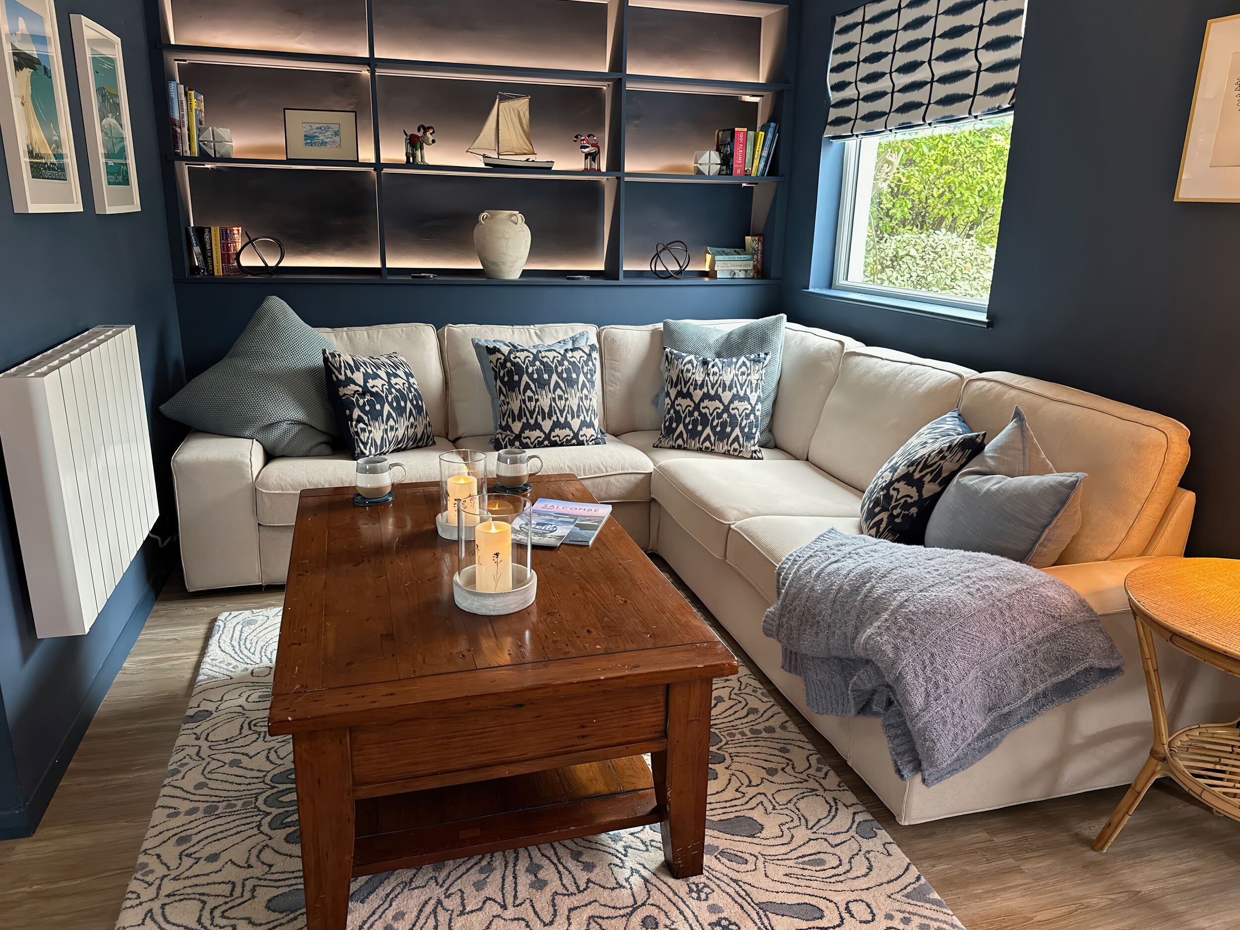 Una sala de estar acogedora con un sofá blanco, cojines estampados, una mesa de centro de madera con velas y una estantería con diversos objetos.