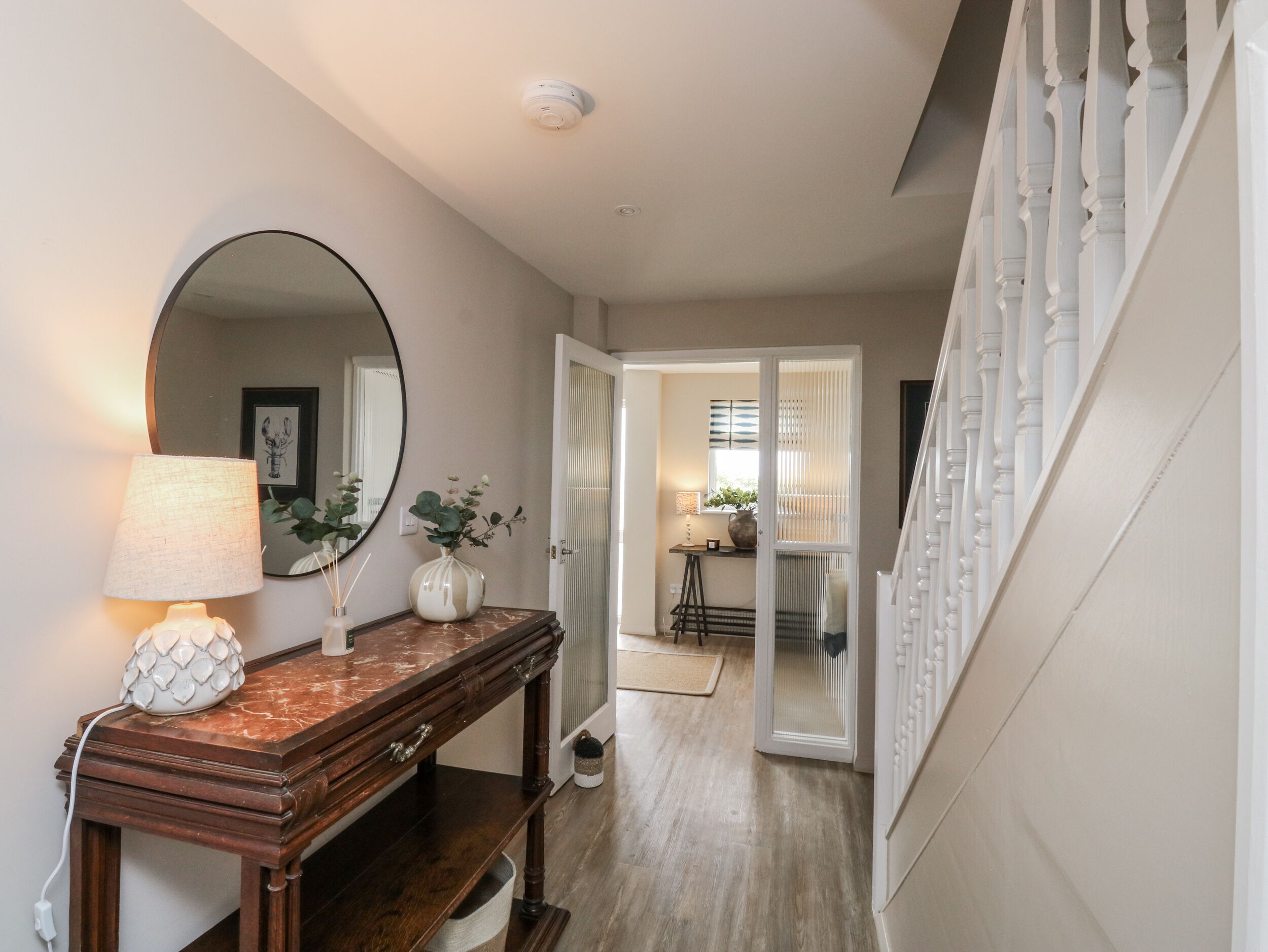 A hallway with a wooden console table, a round mirror, a lamp, and a vase with flowers.