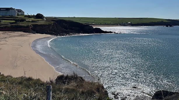 Un paysage côtier avec une plage de sable, une mer bleue et limpide, et une colline herbeuse avec un bâtiment en arrière-plan.