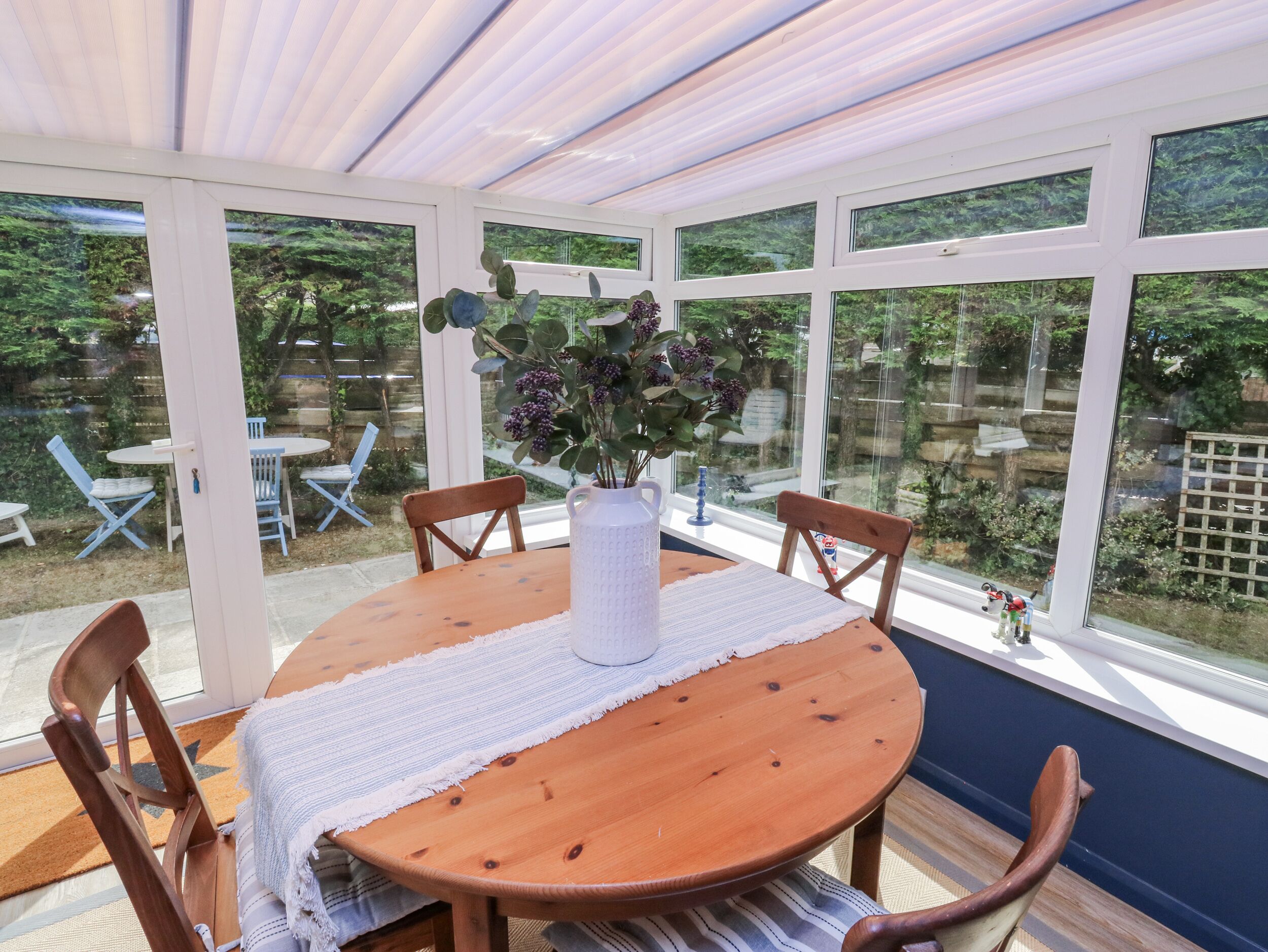 Ein Wintergarten mit Holztisch und -stühlen, einer weißen Vase mit Blumen und Blick auf den Garten durch große Fenster.