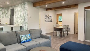 A modern living room with a grey sofa, a blue ottoman, and a dining area with a wooden table and chairs. - Two Cottages on Waterfront Property with Beach Access - Sleeps 10 (Sooke)
